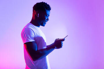 A young black man interacts with his smartphone in a vibrant neon-lit setting. He appears focused and happy, enjoying his online experience and messaging friends.