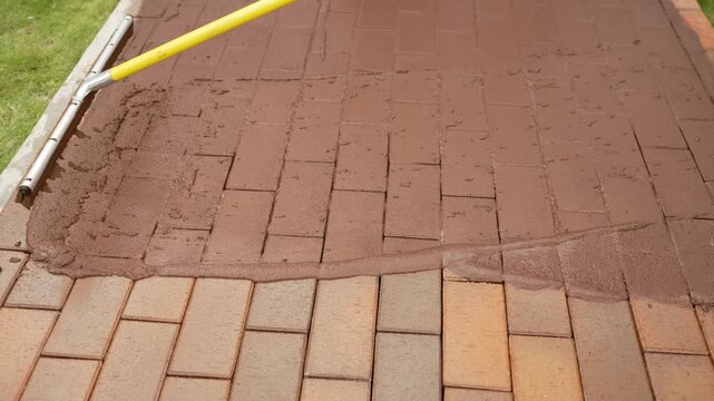 Worker applying a wet grout mixture to seal the joints of a newly laid brick patio using a squeegee tool