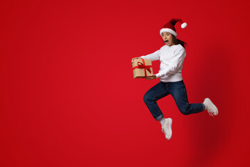 A person wearing a Santa hat jumps in mid-air while holding a gift box. The bright red background...