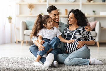 Cheerful middle-eastern family young father, pregnant mother and cute little daughter waiting for baby, sitting on floor in living room, bonding and touching moms big belly, copy space