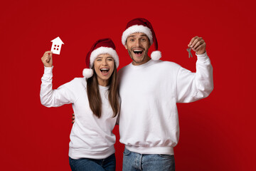 Fototapeta premium A joyful couple wearing Santa hats stands together, excitedly holding a small paper house and keys. Their smiles radiate holiday cheer in front of a vibrant red background.
