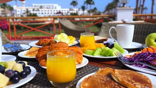 Person enjoying a delicious and nutritious breakfast buffet on a sunny terrace at a luxury hotel, picking a piece of fresh melon with a fork from a plate full of assorted fruits
