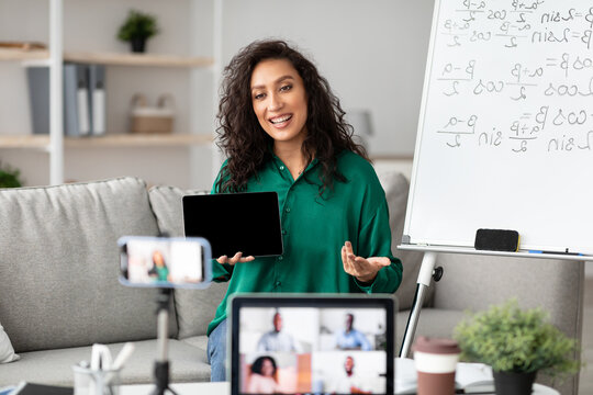 Female math teacher making video conference chat with diverse group of students using laptop and cellphone on tripod, holding showing tablet with empty screen with copy space for mockup, explaining - Powered by Adobe