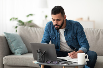 Serious young Arab man using laptop, working remotely, taking notes during remote business meeting from home. Millennial Eastern guy studying online, writing down info. Modern technologies concept