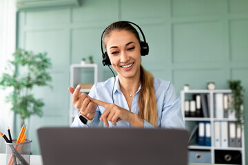 A young female teacher engages in a video conference, explaining concepts to her students. She gestures expressively while using a laptop and wearing headphones in a well-lit room.