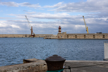 scenic view of a coastal harbor featuring a historic lighthouse, industrial cranes, stone fortifications and calm blue water under a dramatic cloudy sky