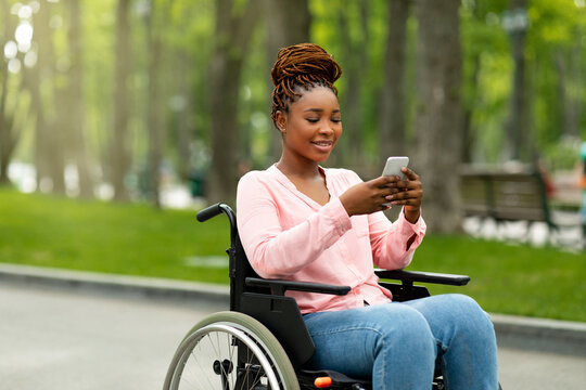 Happy handicapped black woman in wheelchair using smartphone, checking messages, browsing internet at city park. African American disabled lady with mobile device posting in social network outdoors