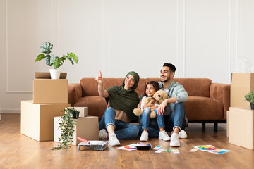 Portrait of smiling muslim family sitting on the floor leaning on couch in new apartment, doing...