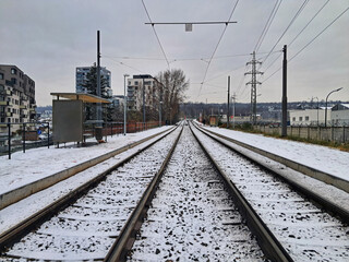 Fototapeta premium Straight tram tracks are covered with a dusting of snow, leading away towards a modern urban landscape and a train station platform in a Prague residential area.