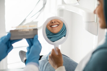Naklejka premium Beautiful Muslim Woman In Hijab Looking At Mirror In Dentists Office, Islamic Female Patient Enjoying Her Beautiful Smile And Teeth After Dental Treatment In Stomatologic Clinic, Selective Focus