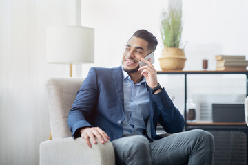 Fototapeta premium Smiling Arab businessman talking on mobile phone at office, sitting in armchair, having pleasant conversation with friend or client. Office employee in formal suit speaking on smartphone indoors