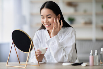 Pretty asian woman in bathrobe applying anti-aging face cream in white jar, sitting at table in...