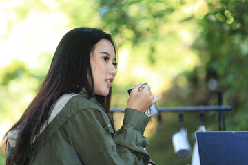 Pretty Young Asian Woman is Sitting Outdoor at a Camping Ground Holding Cup