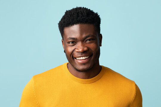 Portrait of handsome young african american guy in stylish yellow jamper posing alone on blue studio background, cheerfully smiling at camera, copy space. Millennials males guys photos concept