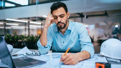 A man is seated at a table in an office space. He is reviewing blueprints and appears to be deep in thought. Tools and a laptop are present on the desk.