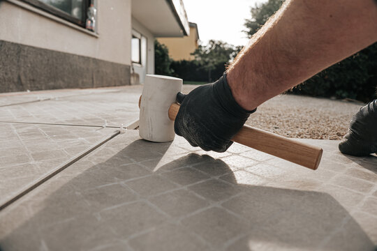 Terrace construction.Tile laying works.Laying Patio Tiles with Rubber Mallet.Person Installing Stone Pavers with Rubber Mallet. construction hammer taps tiles.Hands in black gloves lay tiles on a