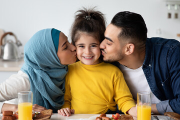 Loving Parents Kissing Their Little Daughter To Both Cheeks While Eating Breakfast In Kitchen,...