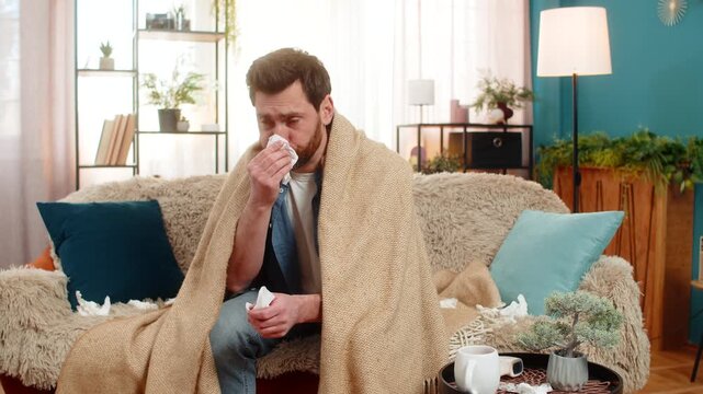 Young sick ill man at home sits on the sofa, wrapped in blanket, sneezing and quickly reaching for tissues. Caucasian guy wipes his nose, looking unwell, possibly suffering from a cold or flu symptoms