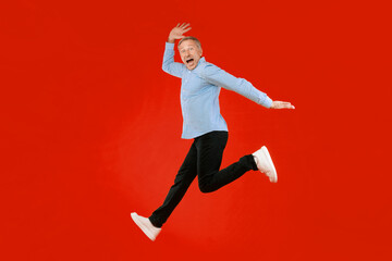 A man with gray hair and a blue checkered shirt jumps in the air with an excited expression. His black pants and white shoes contrast with the bright red background. The scene shows joy and energy.