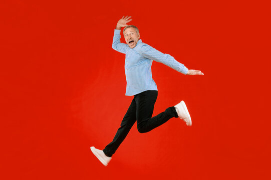 A man with gray hair and a blue checkered shirt jumps in the air with an excited expression. His black pants and white shoes contrast with the bright red background. The scene shows joy and energy.