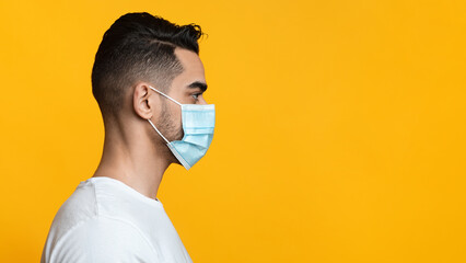 Profile portrait of arab guy with protective face mask over yellow studio background. Middle-eastern young man wearing medical face mask while coronavirus pandemic, looking at copy space, panorama © Prostock-studio