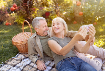 Modern elderly spouses taking selfie on smartphone, having picnic and resting in garden at sunny autumn day, copy space. Senior couple capturing happy moments