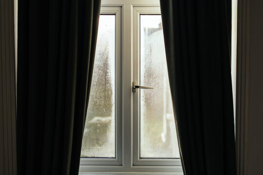 Condensation forming on double-glazed window with closed dark curtains in winter light.