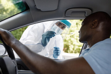Black man sitting inside car, making drive-in pcr rest outdoors. Doctor or nurse in protective suit standing by open auto window, holding swab for covid pcr test, assisting african american driver