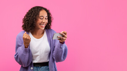 A girl celebrates a happy moment while looking at her smartphone. She has curly hair and is wearing...