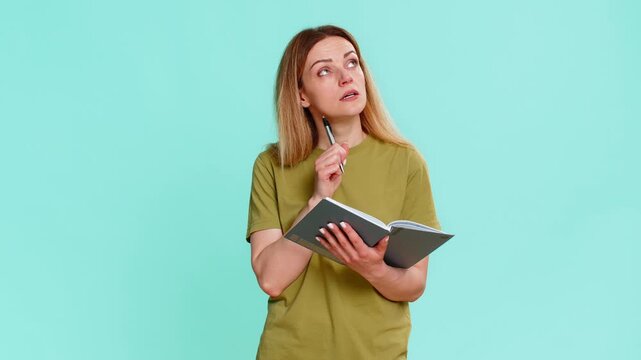Middle-aged woman writes notes in notebook, pauses with thoughtful expression and looks up, reflecting on life direction or next step. Girl on blue background shows deep concentration and planning