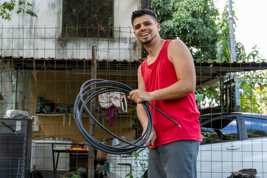 Smiling man wearing a red sleeveless shirt rolling up a black cable in his backyard.