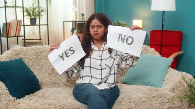 Indian woman at home holds yes and no papers hesitating choosing feeling stressed confused. Arabian girl on sofa looks at camera seeking advice showing doubt decision dilemma anxious mood alert tense.