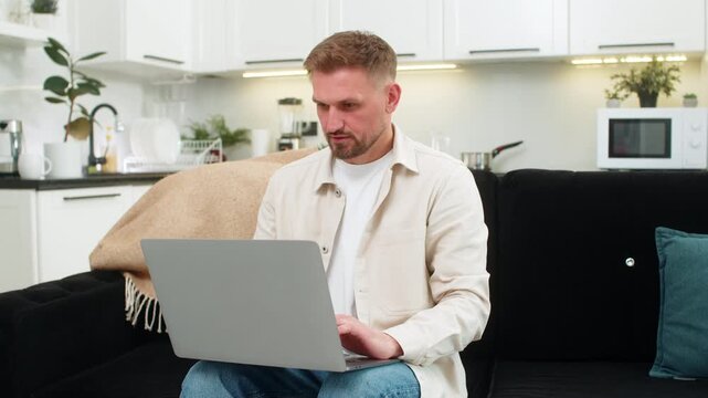 Young man at home opens laptop starts work launches system and types fast with focus and confident smile appears. Caucasian guy on sofa checks tasks writes notes shows productive mood with clear goal.