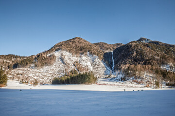 Wide winter view of Zgornje Jezersko in Slovenia with snow covered plain, scattered farm buildings and forested mountain slopes under a clear blue sky.
