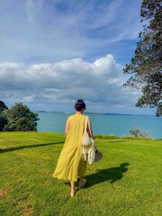 Fototapeta premium A girl in yellow dress is looking at beautiful blue ocean under white clouds and blue sky in New Zealand.
