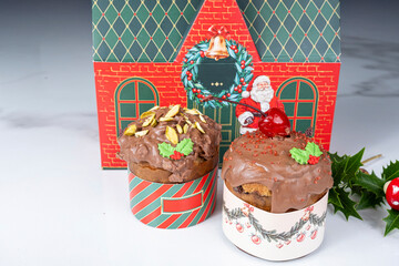 Small panettone cakes with chocolate coating next to a box shaped like a Christmas house_front view.