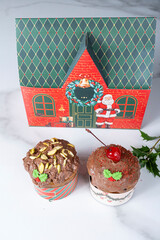 Small panettone cakes with chocolate coating next to a box shaped like a Christmas house_vertical.