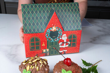 A woman holding a box shaped like a Christmas house next to panettone cakes covered in melted chocolate.