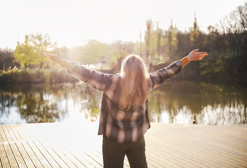Back view woman on wooden pier arms wide open facing lake, autumn sunshine, freedom and gratitude,...