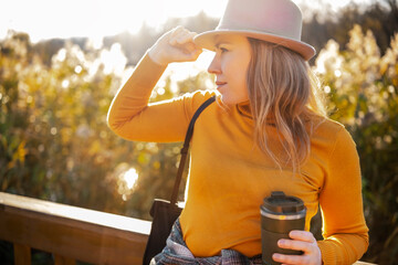 Woman leaning on wooden railing holding hot drink, autumn park near lake, warm casual outfit, slow outdoor walk, relaxed lifestyle and nature mood