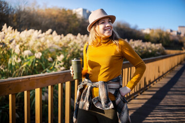 Happy woman smiling on wooden bridge near lake, yellow sweater and hat, autumn sunlight, peaceful...