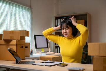 Stressed asian female entrepreneur holds her head in office with boxes, tablet and computer