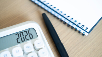 Close-up of calculator displaying 2026 with pen and notebook on wooden desk, representing financial...
