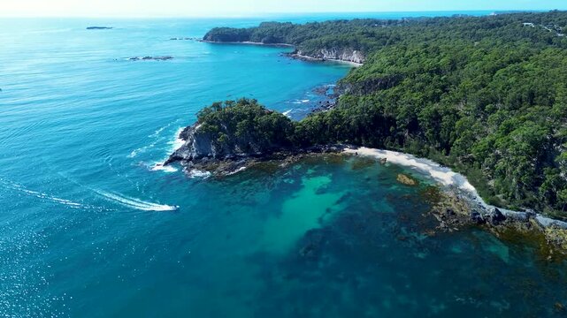 Drone aerial landscape showing boat arriving in sandy ocean cove beach with rocky mountainous headland coastline and forest trees in background of Batemans Bay Eurobodalla Australia nature tourism