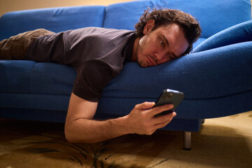 Chilled man on sofa, Casual individual reclining on sofa engrossed with mobile device, Unwinding man sits comfortably on sofa focused on his smartphone amid cozy modern decor