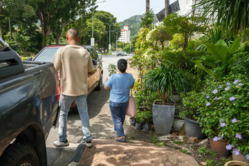 Two people walking along a street with plants and cars under bright sunlight in a city setting
