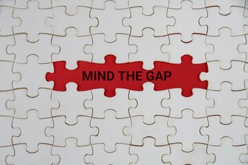 A red puzzle piece stands out amid white pieces, with the words Mind The Gap. It conveys focus on gaps, problem solving, and teamwork toward solutions.