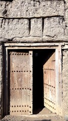 Weathered Clay Wall Reveals Rustic Craftsmanship, Partially Open Heavy Wooden Doors Leading To Shaded Courtyard, Ancient Adobe Doorway With Carved Walls Opens To Secluded Sunny Courtyard Scene