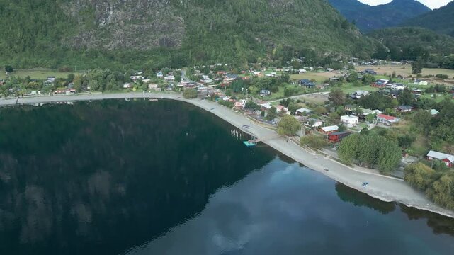 Tilting down drone aerial of Ranco Lake village with houses, sandy beach, pier, dramatic rocky mountains backdrop, and Chilean shoreline