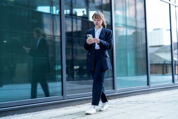 Business professional walks on city sidewalk while using smartphone in front of modern glass building